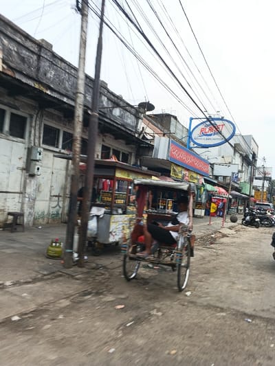 The image depicts a daytime street scene in Bandung City, Indonesia, characterized by urban infrastructure and commercial activity under an overcast sky. In the foreground, a paved but dirty street runs diagonally from bottom left to top right, showing debris and discolored patches. A traditional three-wheeled pedicab (becak) occupies the mid-ground, traveling right to left. A male operator, wearing a white short-sleeved shirt and dark pants, is seated in the driver's position, facing slightly left.
To the left of the pedicab, a portable vendor's stall with a canopy and displayed merchandise (text not fully legible) is positioned on the sidewalk, partially obscured by a dark utility pole. Another utility pole stands prominently in the center foreground. A green LPG tank and a small wooden stool are visible on the sidewalk to the far left.
The left side of the image features a weathered, multi-story building with dark, closed windows and a dilapidated facade. Overhead, numerous black electrical or communication wires crisscross the sky, attached to utility poles.
In the mid-ground to the right, a bright blue oval sign with "PLANET SERVIS & BAN MOTOR" in white text is mounted above a row of commercial establishments. These buildings display various signs in red, yellow, and blue, indicating shops or services. Further down the street, on the right, several motorcycles are parked in a cluster. The street extends into the background, where more buildings and parked vehicles are visible, fading into the distance.