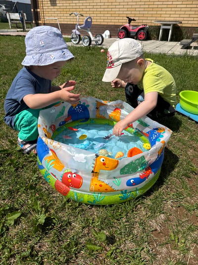 In Zav'yalovo, Russia, two young children are playing in a small, inflatable kiddie pool filled with water and small plastic toys. The child on the left, wearing a blue t-shirt, green pants, and a blue bucket hat with a sailboat pattern, is looking down into the pool with their hands submerged. The child on the right, wearing a yellow t-shirt and a white baseball cap, is also looking into the pool and reaching for a toy. The kiddie pool has a colorful design featuring cartoon animals, including dinosaurs and fish. The scene is set on a sunny day in a grassy backyard, with a brick house and some outdoor toys, including a pink ATV and a purple tricycle, visible in the background.