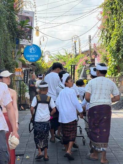 In Ubud District, Indonesia, a group of people, including several young boys dressed in white tops and traditional sarongs, walk down a paved street lined with lush greenery and flowering vines. One boy in the foreground wears a white checked shirt and a dark purple sarong with gold embroidery. To his left, another boy in a white shirt and sunglasses looks off to the side. A woman in a white visor and floral top is visible on the left, and other individuals are seen in the background. Overhead, a tangle of electrical wires crisscrosses the sky. A circular blue sign with the text "SAMATHA SPA" and a woman's silhouette is mounted on a pole. Another sign advertising "LAUNDRY" is also visible. Some of the individuals are pushing a wheeled cart. The overall scene suggests a busy street in a tropical urban environment, possibly during a cultural event or procession.