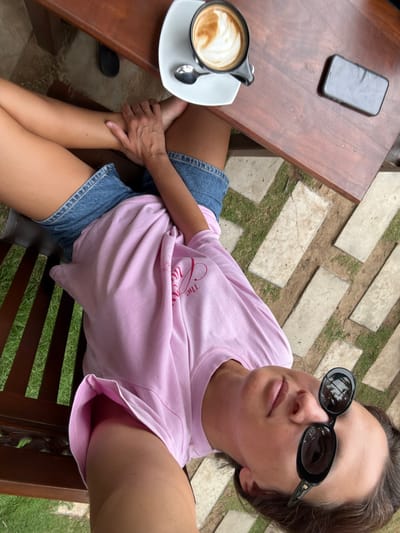 A woman in black sunglasses and a pink t-shirt poses in an outdoor setting. She is seated on a wooden bench with her legs crossed. A wooden table beside her holds a smartphone and a cup of coffee with a spoon. The background includes a stone-paved area with grass growing in between the stones.