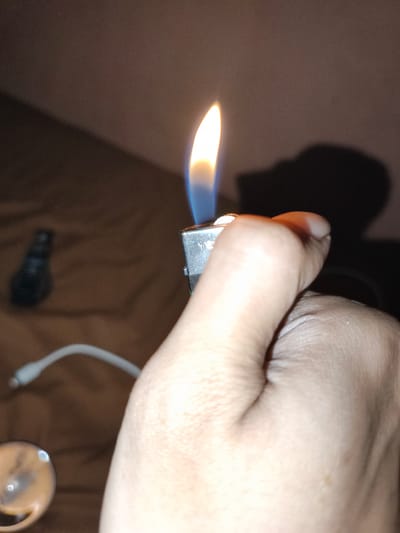 A close-up, indoor photograph shows a human hand holding a lit silver lighter. The flame is mostly yellow, with a blue base. A shadow is cast on the wall behind the hand. Out of focus in the background, on what appears to be a bed or similar surface, is a dark object (possibly a vape pen), a white cable, and what appears to be a small glass pipe. The image was likely taken in Pacet, Indonesia.