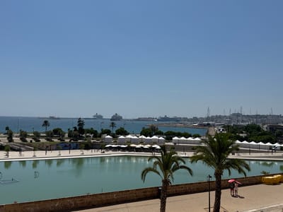 The image captures a wide, elevated view of the waterfront in Palma de Mallorca, Spain. In the foreground, a large rectangular body of turquoise water, likely a reflecting pool or decorative pond, dominates the lower half of the frame. A low stone wall borders the pond. Two palm trees stand to the right of the pond, with a man and woman holding pink umbrellas standing on the paved walkway behind them. Beyond the pond, a paved promenade is visible, with trees, lampposts, and a few white tents set up. In the middle ground, the azure sea stretches out, dotted with several large cruise ships. A long breakwater extends into the water. The background features a cityscape with buildings and a harbor filled with numerous sailboats and yachts. The sky is clear and blue, indicating a sunny day.