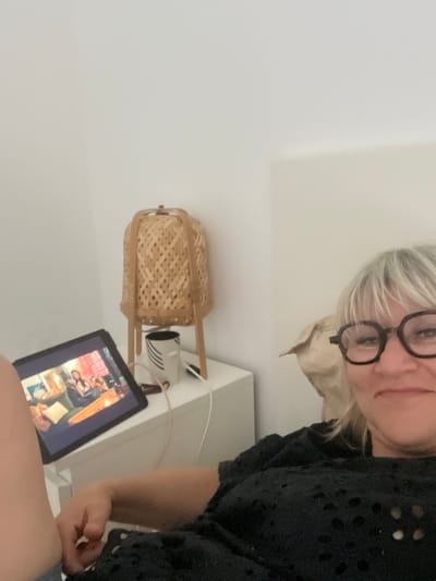 In a bedroom, a woman with blonde hair and glasses is lying on a bed, looking at a tablet computer. She is wearing a black shirt. The tablet is on a nightstand, next to a mug and a lamp with a woven shade. The room is sparsely decorated with a white wall. The image was taken in Chambœuf, France.