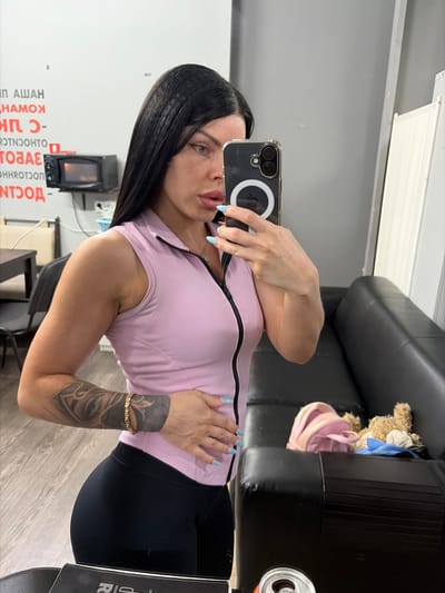 The media file shows a woman taking a mirror selfie in what appears to be a gym or fitness studio, located in Ufa, Russia. The woman has dark hair and is wearing a light pink sleeveless top with a zipper and black leggings. She has light blue nail polish and a tattoo on her left arm. She is holding a smartphone with a ring light attached to it, which she is using to take the photo.
In the background, a black leather couch is visible, with a teddy bear and a pink bag placed on it. To the left of the woman, there is a microwave oven and some signage with text. The lighting suggests it is daytime. The woman's expression is neutral, and she appears to be posing for the photo. The overall setting is clean and modern, consistent with a fitness facility.