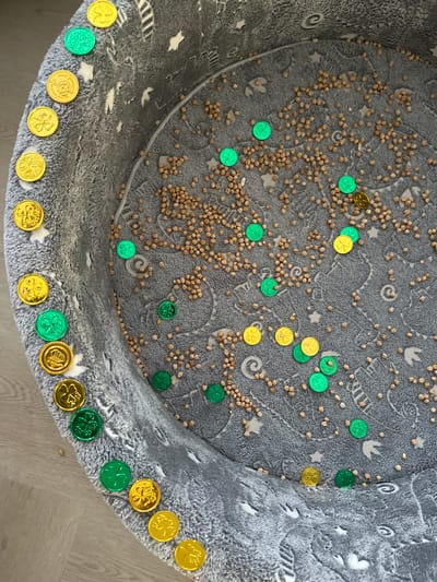 The image shows a grey, circular fabric container filled with dried chickpeas and numerous metallic coins, both green and gold, which depict shamrock designs. The coins are scattered throughout the chickpeas, with a cluster of them arranged along the left edge of the container. The container appears to be a pet bed or a sensory bin for children. The background is a light-colored wooden floor. There are no people or animals visible in the image, nor is there any indication of the location being in Leeds, United Kingdom.