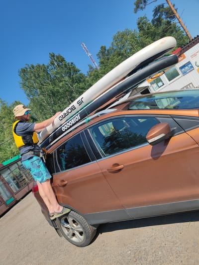An adult male wearing a straw hat, sunglasses, a yellow life vest, and blue patterned shorts stands on the rear tire of a brown SUV, holding onto two large inflatable paddleboards strapped to the roof rack. The paddleboards are white and black, with "JS BOARD" and "SCIROCCO" printed on them. The SUV is parked on a gravel surface, with trees and a building in the background. A red and white communication tower is visible in the sky above the trees. The environment appears to be a sunny day in a rural or suburban area, possibly near a body of water given the paddleboards. The building in the background has signs in Russian, suggesting the location is in Russia.
