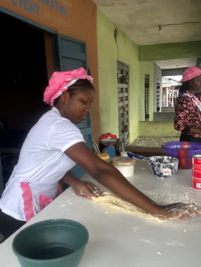 In Badagry, Nigeria, a young woman with braided hair and wearing a pink head covering and a white t-shirt is actively engaged in preparing dough. She is standing at a white surface, possibly a table, which is dusted with flour. Her hands are covered in dough as she works it, her focus intent on the task. Next to her are various objects associated with food preparation, including a blue plastic basin containing a red tin, a white plastic cup, and a blue and white scoop. A large, dark green plastic bowl or basin is also visible.
The scene appears to be outdoors or in an open-air setting, suggested by the building structure in the background. The walls of the building are painted in shades of green and light beige, with windows that have metal grates. The overall impression is one of a functional space, possibly a home or a small business. The light suggests it is daytime. The woman's focused expression indicates concentration on her work. A sign with text is partially visible on a brown surface in the foreground, with the words "EVENT" and "ATION" clearly legible, along with a phone number. The overall atmosphere is one of everyday activity and labor.