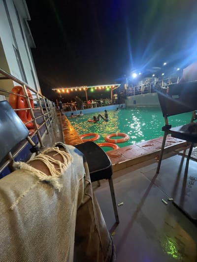 A person sits beside a pool on a boat at night. Ripped denim jeans are visible in the foreground. The pool has several orange life rings, and multiple people swim and stand in the water. The boat is lit with string lights and artificial lights, with part of the deck and upper levels visible. The dark night sky surrounds the boat.