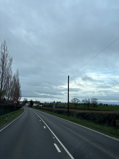The image displays a two-lane asphalt road under an overcast sky in Ruyton-XI-Towns, United Kingdom. The road curves gently to the left in the foreground and features a solid white line marking the left edge and dashed white lines indicating the center and right edge of the carriageway. On the left, a dark fence runs parallel to the road, succeeded by a row of tall, bare deciduous trees. A red and white triangular road sign, indicating a junction, is visible, along with multiple small white bollards topped with red. Further down the road, faint pink blossoms are present among trees, and several residential buildings are discernible. Two large white vehicles, consistent with lorries or trucks, are observed in the distance, proceeding towards the viewer. On the right side of the road, a green verge borders a dense hedge, beyond which a brown ploughed field and a distinct green field are visible. A prominent dark utility pole stands on the right, from which a single dark wire extends diagonally upwards across the sky. A row of bare deciduous trees lines the distant field edge, and faint hills are visible on the horizon. The sky is characterized by multiple layers of grey overcast clouds with some lighter patches. No individuals are present in the scene.