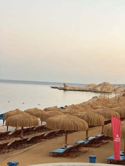 This image depicts a serene beach scene at Park Regency Resort in Sharm El Sheikh, Egypt. The foreground is dominated by rows of neatly arranged beach chairs and thatched umbrellas, casting shadows on the sandy shore. The calm, tranquil sea stretches to the horizon, with a rocky outcrop jutting into the water on the right side of the frame. The sky is overcast with a soft, diffused light, suggesting either early morning or late evening. A red flag with the "Park Regency Resort" logo is prominently displayed on the right, indicating the resort's presence. Several red buoys are visible in the shallow water, marking a swimming area. The overall impression is one of peace and relaxation, characteristic of a resort destination.