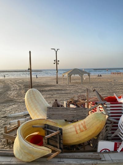A beach scene shows the shore on the left and sand in the middle. Various man-made structures are present on the beach, including gazebos, utility poles, and a lifeguard station. Near the top right, stacked chairs are visible. The focus is on a large, yellow object resembling a banana or banana peel. This object sits on a wooden structure, with a red bucket visible inside its curved opening.