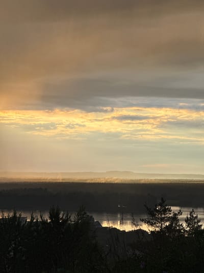 The image captures a serene and atmospheric landscape, likely at sunrise or sunset, over a large body of water in the Прикамский region of Russia. The sky dominates the left side (upper portion when oriented correctly), showcasing a dramatic display of golden and warm orange light filtering through scattered clouds. These clouds are brightly illuminated, creating long, streaky formations against a softer, hazy background.
A faint, light-toned strip of land, possibly distant hills or a far shoreline, is visible across the middle of the image, appearing somewhat muted by atmospheric haze. Below this, the expansive body of water (likely the Kama River or a reservoir) reflects the sky's radiant light, showing shimmering streaks of gold and silver on its surface.
In the foreground, on the right side of the image, dark, silhouetted trees and bushes add depth and contrast to the scene. Their intricate branches are sharply defined against the glowing water and distant horizon. No people or visible text are present. The overall impression is one of peaceful natural beauty, with the warm, diffused light creating a tranquil and majestic ambiance.