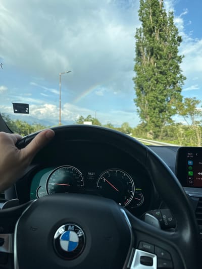 The image, taken from inside a car in Novo Delchevo, Bulgaria, captures a driver's perspective on a road. A person's right hand is visible on the BMW steering wheel in the foreground, indicating an active driving scene. The car's dashboard is prominent, featuring a speedometer reaching 260 km/h, with the needle resting near 60 km/h, and a tachometer showing RPMs. A digital display on the dashboard indicates the time as 18:21 (6:21 PM) and an outside temperature of +23.0°C. Other visible controls on the steering wheel and stalk include "MODE" and "AUTO."
Through the windshield, a road lined with lush green trees is visible under a sky filled with scattered clouds. A notable detail in the sky is a faint but distinct rainbow arcing across the upper left. A light pole or structure also protrudes from the left side of the frame. On the car's dashboard, a smartphone displaying a similar time (18:21), "5G" connectivity, and various app icons (including phone, messages, WhatsApp, Spotify) is mounted. Another time stamp on the phone's screen shows "2:15" with "Done." A small, dark rectangular sticker with some illegible text is affixed to the lower left of the windshield. The overall impression is a calm drive, possibly after rain given the rainbow, during the early evening.
