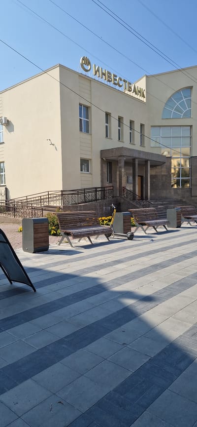 The image shows a sunny day in front of a light yellow building with a sign that reads "ИНВЕСТБАНК" in gold letters. The building has several windows and a rounded arched window above the entrance. In front of the building, there are several wooden benches and two dark gray trash bins. A yellow electric scooter is parked next to one of the benches. The ground is paved with gray and dark gray rectangular tiles arranged in a striped pattern. A shadow is cast across the tiles from the left side of the frame. A ramp with a black railing leads up to the building. In the background, there are power lines stretching across a clear blue sky. A white object, possibly a bird, is seen flying near the building. The scene appears to be a public area in the city of Miass, Russia.