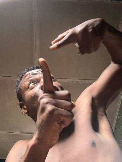 This media file is a close-up, low-angle shot of a young Black man in what appears to be a bathroom or a similar tiled indoor space, likely in Ikorodu, Nigeria. The man is shirtless, and the lighting is dim, suggesting it might be nighttime or a poorly lit interior.
The man's head is tilted, and his eyes are looking directly at the camera, conveying a serious or intense emotion. He is making a hand gesture with both hands. His right hand is held up, with fingers extended in a gesture that could be interpreted in various ways, including a sign of aggression or a stylistic pose. His left hand is also raised and positioned near his face, with fingers bent. The focus is primarily on his upper body, face, and hands, with the background consisting of tiled walls.
Notable details include the texture of his skin, the definition of his muscles, and the lines on his forehead, which are accentuated by the lighting. His hair is short and textured. There is no visible text in the image. The overall impression is one of a candid, possibly staged, moment captured with dramatic lighting.