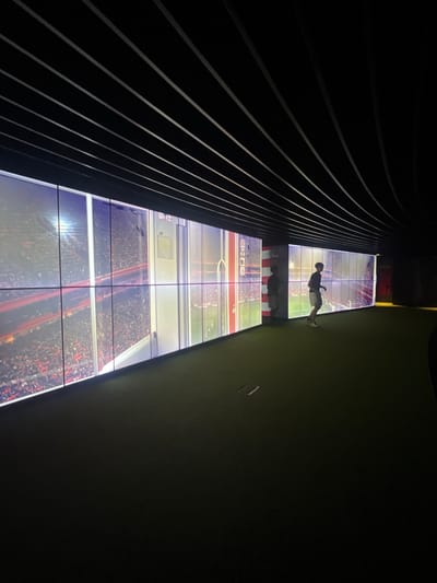 The image shows an immersive exhibition space with a large, continuous video screen displaying a sports stadium filled with spectators. A young man is walking on the green carpeted floor in front of the screen. The space has a dark ceiling with parallel, linear lighting elements and a curved wall on the right side. The video screen covers the majority of the left and central wall, showcasing a bright, detailed image of a stadium. The man, silhouetted against the screen, is wearing a dark t-shirt and light-colored shorts, with white sneakers. His posture suggests a casual walk. The environment is dimly lit, with the primary illumination coming from the video display. This exhibit is located at the Athletic Club Museum of Bilbao, in the city of Bilbao, Spain, as indicated by the museum's context.