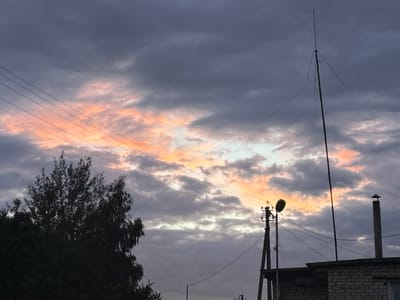 The image depicts a twilight scene with a dramatic sky in the city of Забалоцкі сельскі Савет, Belarus. The sky is filled with dark, heavy clouds interspersed with patches of bright orange and pink hues, suggesting the last rays of sunlight. Several power lines crisscross the frame diagonally from the upper left. In the foreground, silhouetted against the sky, is the dark foliage of a large tree on the left side. To the right, there are man-made structures including a utility pole with a light fixture, a tall slender antenna, and a brick building with a chimney. The overall impression is one of a slightly industrial or suburban landscape under a moody sky. No people are visible in the image.