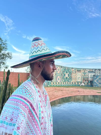 A man wearing a colorful sombrero and a white poncho with pink and turquoise patterns stands in the foreground, facing to the right. Behind him, a decorative wall with geometric patterns is visible, along with a body of water and a few cacti. In the background, a white animal, possibly a goat, grazes. The sky is blue with scattered clouds. The location is likely a resort or tourist attraction in Mui Ne, Vietnam, given the presence of cacti and decorative elements common in such establishments, and the style of the man's attire is reminiscent of traditional Mexican cultural dress, possibly for a themed event or photoshoot.