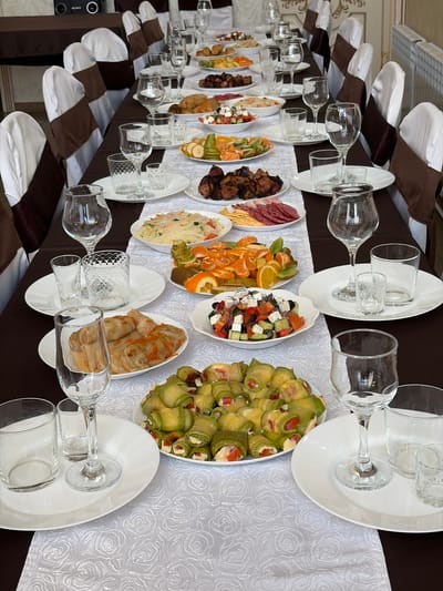 A long banquet table laden with various dishes and glassware is seen from an elevated, side angle. The table is covered with a white tablecloth featuring a subtle rose pattern. Each place setting includes a large white dinner plate topped with a smaller plate and two glasses – a wine glass and a smaller drinking glass. Dishes of food are spaced along the table's length, including zucchini rolls, cabbage rolls, mixed fruit (oranges and kiwi), salad, charcuterie (salami, meat, cheese), and roasted meats. Chairs along the table are adorned with white covers and brown sashes. The location is in Neftekamsk, Russia.