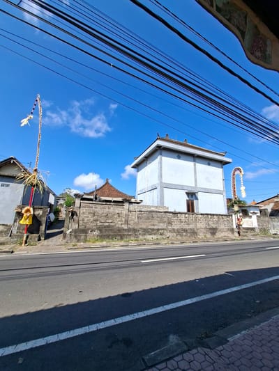 The image captures a street scene in Blahbatuh, Indonesia, under a clear blue sky with scattered clouds. A paved road runs horizontally across the foreground, with white lane markings visible. To the left of the road is a sidewalk with hexagonal paving stones. Behind the sidewalk is a low, concrete wall constructed from cinder blocks.
On the left side of the image, beyond the wall, is a gray house. In front of this house, a tall, thin pole with decorative elements is planted vertically. This pole features a red section near its base, with straw-like decorations and a yellow object suspended from it. Further back, another house with a red tiled roof is partially visible.
To the right of the first house, a more prominent, multi-story building with a light blue facade stands behind the cinder block wall. This building has a traditional pitched roof with decorative elements. Another decorative pole, similar to the one on the left but taller and more elaborate with white and red elements, stands near this building. A smaller, white structure is situated to the right of the blue building.
The environment is characterized by dense overhead power lines that stretch across the top of the frame. The buildings appear to be residential or commercial structures typical of the region. The overall impression is of a quiet street in a tropical climate, possibly with some form of local celebration or decoration.