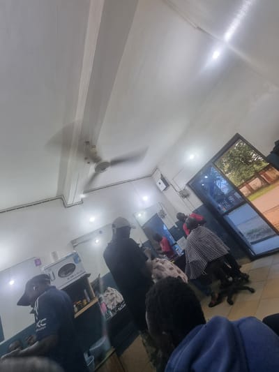 The image shows the interior of a barber shop in Gwong, Nigeria. Several individuals are present. In the foreground, a person wearing a blue hoodie is facing away from the camera. To the left, a person wearing a blue shirt with a white logo and a black cap is standing. Further back, a person in a black t-shirt and a white cap is standing, and another person in a red shirt is seated. A person wearing a black and white striped smock is also seated. A ceiling fan is visible on the ceiling. A large glass door reflects the outside environment, showing trees and a building. Shelves with various items, including a white box labeled "TOWEL WARMER UV STERILIZER," are mounted on the walls. The lighting appears to be from overhead fixtures and possibly natural light from the door.