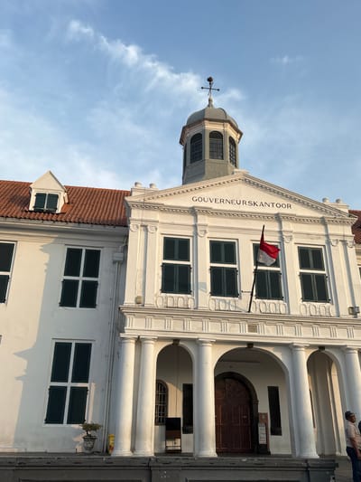 The image shows the exterior of the Fatahillah Square (Kota Tua) building in West Jakarta, Indonesia. The building is white with a red tiled roof and features multiple windows with dark green shutters. Above the entrance, the text "GOUVERNEURSKANTOOR" is inscribed. An Indonesian flag, red and white, is flying from a flagpole attached to the facade. The sky above is blue with some wispy white clouds. In the lower right corner, a person wearing a cap and light-colored shirt is visible. The building's architecture is classical, with columns supporting an arched entrance.