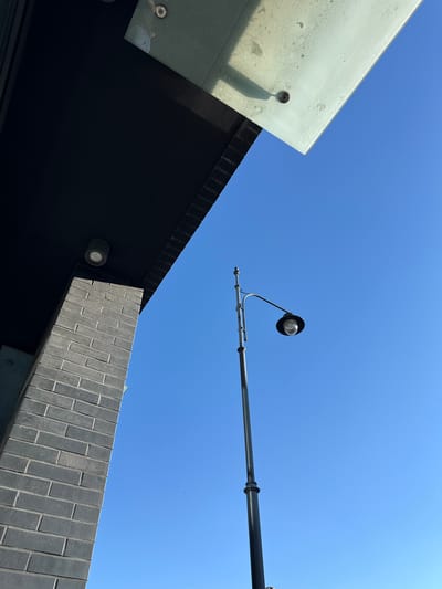 This low-angle shot captures a bright, clear blue sky above a section of modern architecture. The architectural elements include a large, translucent glass overhang, a dark, angular structure that appears to be part of a building facade, and a wall clad in dark grey brickwork. A modern street lamp with a black hood and a clear bulb extends into the frame from the right, its metallic arm also visible. On the underside of the glass overhang, a small, dark surveillance camera is mounted, casting a distinct shadow. Another circular light fixture is visible on the dark facade below. The scene suggests an outdoor urban setting during the daytime, likely sunny, given the sharp shadows. The architecture suggests a contemporary design, possibly a commercial building or public space. No people are visible. No text is discernible in the image. The context provided places this scene in Łódź, Poland.