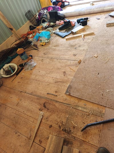 The image depicts a cluttered wooden floor inside a structure, possibly a shed or workshop, in Islambaevo, Russia. The floorboards are aged and dusty, covered with sawdust and scattered debris. Various tools and objects are strewn across the surface, including a drill, extension cords, a metal cage, a can of paint, a small blue plastic container, a tape measure, a white bowl filled with fasteners, and a pair of black athletic shoes. In the background, a chainsaw and a large rolled-up object are visible. The overall impression is one of disarray and ongoing construction or repair work.