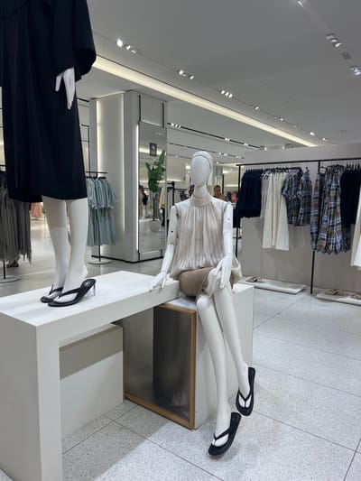 A retail store in Beşiktaş, Turkey, features mannequins displaying clothing and footwear. A mannequin on the left is dressed in a black dress and black flip-flops with clear heels, standing on a white platform. To the right, a seated mannequin wears a light-colored, sleeveless top with a gathered neckline and a neutral-colored skirt, also accessorized with black flip-flops. In the background, clothing racks display various garments, including patterned shirts and solid-colored items. The store's interior has a modern design with white walls, a light-colored tiled floor, and integrated lighting.