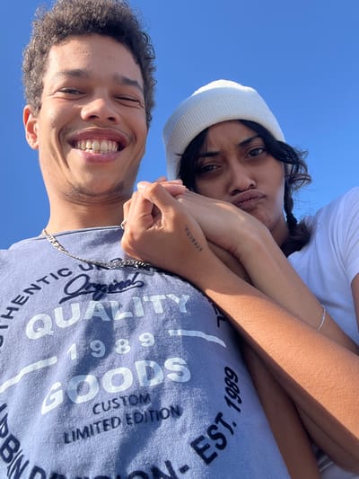 A selfie-style photo shows a man and a woman with a clear blue sky as the background. The man is in the foreground, looking up and smiling widely. He has short curly hair and is wearing a light blue T-shirt with the words "AUTHENTIC UNRE QUALITY GOODS CUSTOM LIMITED EDITION" printed on it. A silver chain is visible around his neck. The woman is in the background, looking at the camera with her lips pursed in a playful expression. She is wearing a white beanie and a white T-shirt. Her arms are crossed over the man's chest, and a small tattoo that reads "breathe." is visible on her arm. The photo appears to be taken from a low angle, suggesting they are looking down at the camera. There is no information to confirm the image was taken in Mitchells Plain, South Africa.