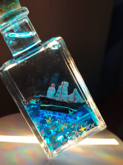 A tilted rectangular glass bottle, approximately three-quarters full, rests on a light-colored, reflective surface. The bottle is sealed with a natural cork stopper. Inside, a miniature dark-hulled ship with three light-colored sails (white with faint red markings) floats above a vibrant blue liquid. The blue liquid contains numerous iridescent star-shaped glitter particles, reflecting a multi-spectrum of colors including gold, green, and pink. A clear liquid layer is visible in the neck of the bottle, emitting a faint blue luminescence. Direct illumination from an external light source casts a prominent horizontal beam of white light and a distinct rainbow prism effect onto the surface beneath the bottle. The background is uniformly dark and out of focus.