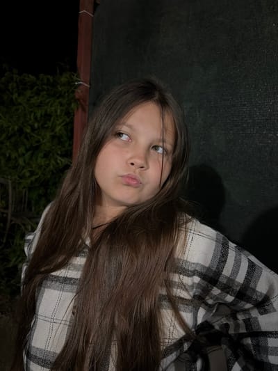 A young girl with long, brown hair and fair skin is posing for a selfie. She is wearing a black and white plaid shirt. The background is dark with some green foliage visible at the bottom left and a dark, possibly textured, surface behind her. The location is unspecified.