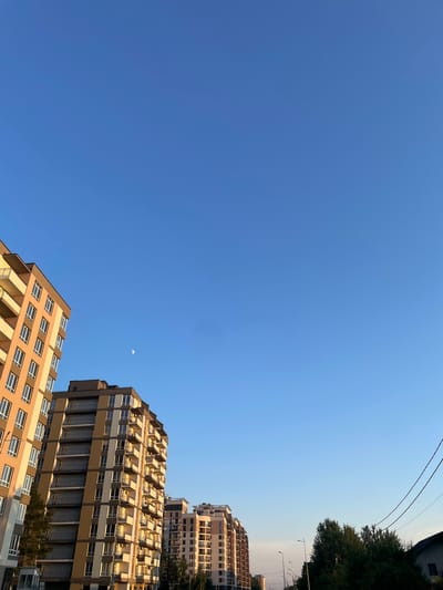 The image shows a clear blue sky with the moon visible as a small, crescent shape in the upper center. Below the sky are several modern buildings, appearing to be apartment complexes, with many windows and balconies. The buildings are angled upwards, suggesting the photo was taken from a low perspective. On the right side of the image, a dense green tree canopy is visible, along with streetlights and power lines. The lighting suggests it is daytime, likely late afternoon or early morning due to the warm light illuminating the buildings and the clear sky. The scene appears to be an urban or suburban environment in Brovary, Ukraine. No people are visible in the image.