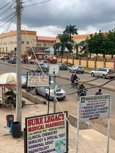 The image shows a street scene in Akure, Nigeria, at the coordinates 7.241 latitude and 5.195 longitude. The street is lined with buildings and palm trees. There are several vehicles parked along the street and some in motion. There are people walking on the sidewalk. In the foreground, there are two signs, one for "MAPET OPTICAL SERVICES & EYE CARE" and one for "SURE LEGACY MEDICAL DIAGNOSIS." The sign for "SURE LEGACY MEDICAL DIAGNOSIS" advertises "ALL MEDICAL TEST" including "CLINICAL CHEMISTRY," "SEROLOGY & SPECIAL TEST," "HORMONAL ASSAY," and "HAEMATOLOGY." The sign for "MAPET OPTICAL SERVICES & EYE CARE" includes a phone number and the address "137, Hospital Road, Akure". The street is paved and has a curb along the side of the road. There is a trash can on the sidewalk. The sky is overcast.