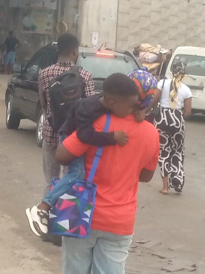 This image depicts a daytime street scene in Badore, Nigeria. In the foreground, a person wearing a red t-shirt and light-wash blue jeans carries a child on their back. The child, dressed in a dark long-sleeved shirt and blue jeans, rests their head on the adult's shoulder. A multicolored shoulder bag, featuring geometric patterns in blue, pink, white, and green with a blue strap, hangs from the child's left shoulder. The child's right foot, clad in a white and black sneaker, is visible. Directly behind the pair, a male individual wears a plaid shirt (red, black, and white pattern), light trousers, and a black backpack. Behind him, a black SUV with its rear brake light illuminated is partially visible. To the right, another individual, possibly female, wears a white t-shirt, patterned black and white pants, and a yellow and brown patterned headscarf, walking away barefoot. A white van, loaded with items on its roof, is parked behind this individual. Another person with a blue and red patterned head covering is partially visible between the carried child and the white van. The street surface is light-colored and appears to be a mix of paved and unpaved areas with some uneven texture. Buildings with concrete or stucco facades are visible in the background, one displaying a poster.