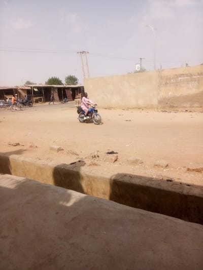 An outdoor scene in Tudun Murtala, Nigeria, depicts a dusty, unpaved reddish-brown road under a hazy, light-blue sky. In the immediate foreground, a concrete structure, likely a drainage ditch or curb, extends horizontally, casting a prominent shadow. An adult male, wearing a light pink traditional garment, operates a dark blue motorcycle, riding across the midground from left to right.
To the left background, a series of open-fronted market stalls or shops constructed with wooden frames and corrugated metal or tarp roofs line the roadside. Several individuals are present near these stalls: one person in an orange shirt stands beside a light-colored bicycle, another person in a dark shirt is positioned near a parked dark motorcycle, and a third individual in a dark reddish garment walks on the road, moving towards the left.
The right background features a tall, plain light-brown colored wall, possibly made of concrete or mud brick. Above this wall, a modern street light on a tall metallic pole is visible. A wooden utility pole with electrical wires stands behind the market stalls on the left. A small cluster of green trees is visible further in the background, behind the stalls. The ground throughout the scene is uneven and scattered with minor debris.