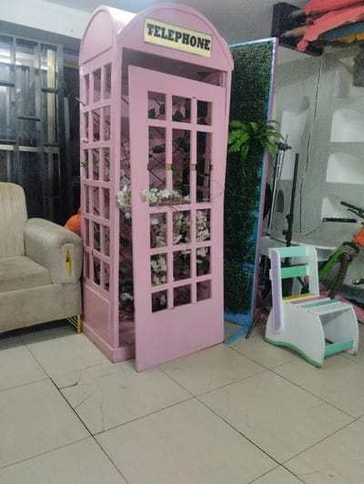 A pale pink telephone booth stands with its door ajar, adorned with white flowers and greenery inside. Next to it, a short, pastel-colored step stool sits on a tiled floor. In the background, there's a green plant wall, a cream-colored armchair with gold accents, and a bicycle partially visible. Mannequins and a metallic structure add depth to the cluttered space.