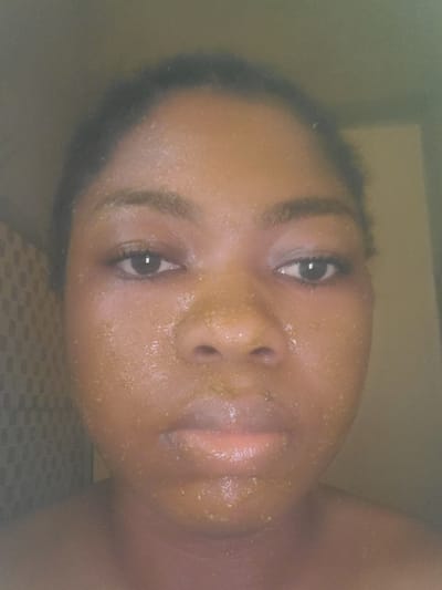 The scene is set in Kakau, Nigeria. A young adult individual with dark skin is depicted in a close-up portrait, facing directly towards the viewer. The person's face is thoroughly covered with a granular, yellowish-brown substance, consistent with a facial mask or scrub, exhibiting small, distinct particles across the forehead, cheeks, nose, and chin. Dark hair is pulled back from the face. The individual has dark, defined eyebrows, and dark eyes that engage directly with the camera. The lips are full and display a natural, moisturized appearance. The shoulders and neck are visible, suggesting the person is bare-chested or wearing a strapless top. The background on the left features a tiled wall with a dark, square grid pattern. The right side of the background is a lighter, uniformly colored wall or surface. The lighting is soft and appears to originate from the front, illuminating the face.