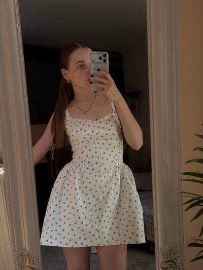 A young woman with auburn hair pulled back in a ponytail, wearing a white and blue floral print dress and a gold necklace, poses for a mirror selfie. She has on multiple earrings and a ring on her right hand. A brown purse is slung over her left shoulder. She is standing in front of a large ornate mirror. The background appears to be an indoor residential setting with a dark wall, a bed with a checkered blanket, and a floor lamp visible. There is a hint of a plant on the right side of the frame. No specific location within Gorzow, Poland can be identified from the image.