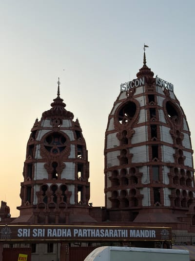 The media file displays two ornate, domed structures with a stark white and reddish-brown color scheme, likely temples. The buildings feature intricate architectural details, including arched openings and decorative elements. A sign attached to the rightmost building reads "SRI SRI RADHA PARTHASARATHI MANDIR ISKCON DELHI", indicating the location as the ISKCON temple in Delhi, India. The structures are viewed from below, emphasizing their height and grandeur.

In the sky above the top structure, a small bird is visible, and a slender spire crowns the building. The overall scene is captured against a pale, overcast sky, suggesting it might be early morning or late afternoon. The lighting is soft and diffused, with a hint of warmth in the sky near the horizon, possibly indicating the golden hour. The perspective is rotated, with the buildings appearing upside down. The overall mood is serene and awe-inspiring, highlighting the spiritual and architectural significance of the location.