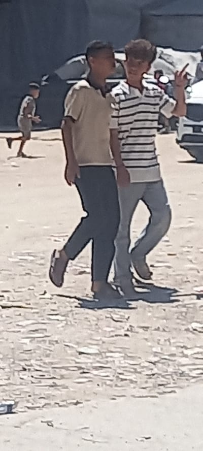 This rotated image, taken in Khan Yunis, Palestinian Territories, captures an outdoor scene during bright daylight.
Three individuals are visible. In the foreground, a person is seen from the back and side, wearing a light brown or tan short-sleeved t-shirt and dark trousers. Their head is turned towards another person standing behind and slightly to their left. This second individual wears a striped short-sleeved collared shirt (white and dark stripes) and light grey pants. They are making a 'V' sign or peace gesture with two fingers of their left hand raised. A third person is partially visible in the bottom left, appearing to be sitting or crouching on a dark surface, wearing light-colored clothing.
The setting is an open outdoor area, with a light-colored, heavily textured ground surface on the right side of the frame, possibly indicating dry earth, sand, or rough concrete. The background on the left is dark and blurred, suggesting a wall or shaded structure. The bright, clear lighting indicates a sunny daytime. The individuals appear to be casually gathered or interacting. No visible text is present in the image.