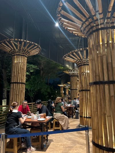 In North Jakarta, Indonesia, a dimly lit outdoor seating area of a restaurant features a distinctive design with large bamboo pillars supporting a corrugated metal roof. Several tables are occupied by groups of people dining. In the foreground, a woman wearing a black shirt and jeans sits at a table, looking at her phone. Across from her, a man in a red shirt is also seated at the table. To their right, a young man wearing a black shirt and glasses is engaged with his phone. Further back, other patrons are visible, some interacting with each other and some looking at their phones. The environment is characterized by the warm glow of lights reflecting off the bamboo structures, creating an intimate atmosphere. Greenery is visible in the background, suggesting a natural or garden-like setting. A blue ribbon barrier is present in the foreground, indicating a designated walkway or queue area.
