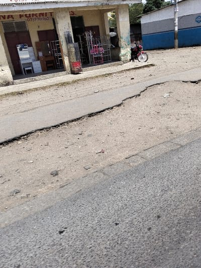 The scene depicts a daylight street view in Mkomani ward, Kenya. In the foreground, a degraded asphalt road surface shows a prominent, irregular longitudinal crack or separation. Adjacent to the road is a dirt-covered shoulder. A concrete sidewalk runs parallel to the dirt shoulder.
On the left, a single-story commercial building with a light-colored facade is visible. The facade displays partial text, "NA FURNIT," and a phone number, "0739701960," indicating a furniture business. Two doorways are present: the left doorway reveals a white vanity table with a mirror and a white stool, alongside darker furniture items; the right doorway displays multiple metal display racks, some painted pink. A tall, red and black cylindrical object stands on the sidewalk in front of this building.
To the right, partially obscured by foliage and the first building, is another single-story structure. This building features a distinctive paint scheme with a blue horizontal band along the lower portion and a lighter band above it, bearing a visible logo.
A single red motorcycle with chrome accents is parked on the dirt shoulder between the sidewalk and the road, positioned in front of the second building and oriented to the left. No individuals or active interactions are present in the image. The background includes visible green foliage and an overcast or brightly lit sky.