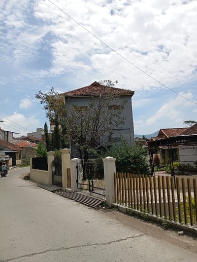 The scene is a residential street in Kecamatan Kutawaringin, Indonesia (latitude: -6.995, longitude: 107.539). There is a two-story house with a gray facade and a red tile roof. The house is surrounded by a black metal fence and a low wall. There is a small gate on the left side of the house. The house is set back from the street, which is paved in gray concrete. On the right side of the street, there is a fence made of vertical wooden slats. Behind the fence, there is a second house with a brown tile roof and a covered patio. There is a green lawn in front of the second house. The sky is blue with white clouds. There is a lone motorcycle parked on the left side of the street, indicating that there may be a resident nearby. The scene is quiet and peaceful, with no people or animals in sight.