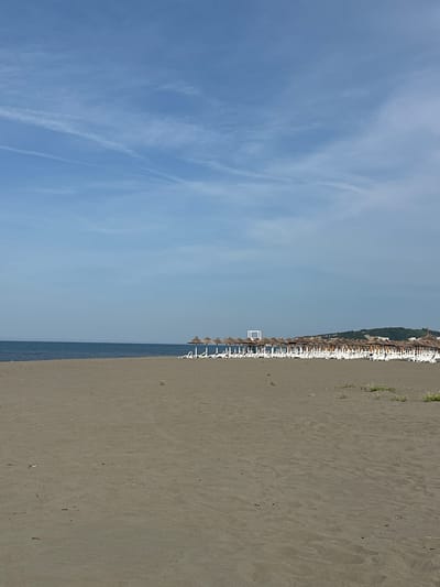 The image depicts a beach scene in Donji Štoj, Montenegro. The scene includes a wide sandy beach on the right, the Adriatic Sea in the center, and a predominantly clear sky with some clouds on the left. A row of beach umbrellas and chairs lines the edge of the beach, near the water. The sand is a light gray or tan color. The water appears calm.