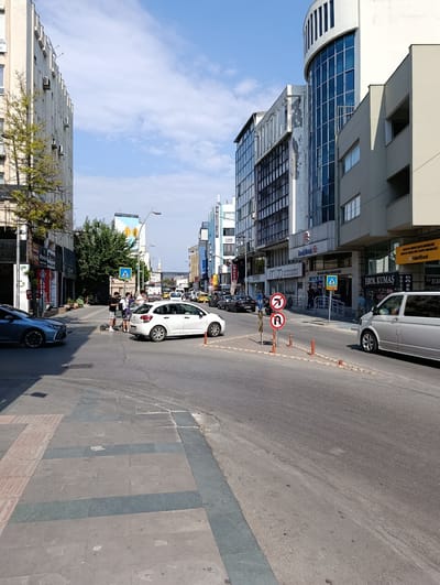 The image captures a street scene in Antalya, Turkey, under a partly cloudy sky. The perspective is from street level, looking down a moderately busy urban road lined with multi-story buildings. On the left, a building with numerous air conditioning units is visible, partially obscured by a tree. The right side of the street features modern architecture, including a building with a curved glass facade. Several cars, including a silver van and a white hatchback, are present on the road. Pedestrians, including a group of young individuals, are seen near a crosswalk. Traffic signs, including those indicating no right turn and a U-turn, are posted. The ground is paved with asphalt on the road and a patterned stone on the sidewalk. The overall impression is of a typical day in a commercial district of Antalya.