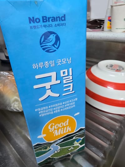 A close-up, eye-level shot captures a light blue carton of "No Brand Good Milk" positioned on a stainless steel sink. The carton, which is the primary subject, features Korean text and a bird logo. To the right of the carton, a white ceramic bowl with red stripes is partially visible. The background is blurred, showing a kitchen counter with various items, including bottles and what appears to be packaging. The lighting is natural and appears to come from above. The scene is likely set in a residential kitchen.