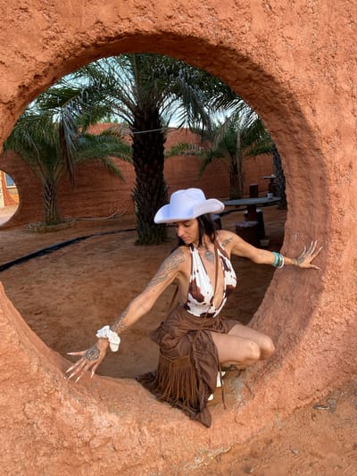 A woman wearing a white cowboy hat and a cow-print halter top is crouched inside a circular, rough-textured, terracotta-colored opening in a wall. She has long, pointed fingernails, tattoos on her arms and legs, and is wearing a brown skirt with fringe. She is leaning with her right hand on the wall and her left hand is extended towards the ground. In the background, palm trees and outdoor seating areas are visible under a blue sky, suggesting a desert or arid landscape, possibly within the city of Mui Ne, Vietnam, known for its sand dunes.