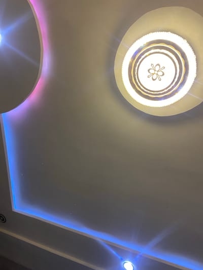 The image shows a modern ceiling with recessed lighting and a decorative chandelier. The ceiling is a neutral color, likely white or off-white, and features a stepped design with illuminated edges. A main chandelier with multiple concentric rings and a floral-like pattern in the center is prominently featured on the left side of the image. This chandelier emits a warm, bright light.
Along the right side of the ceiling, a strip of blue LED lighting creates a vibrant glow, outlining a recessed area. Towards the bottom left, another curved recess in the ceiling is illuminated with a pinkish-purple light, contrasting with the blue light on the right. The overall impression is one of contemporary interior design, likely in a home or a commercial space. The lighting suggests it could be evening or night, or simply that the artificial lighting is the primary illumination source. No people are visible in the image. The location context of Giginyu, Nigeria, does not provide specific visual cues within the image itself. There is no visible text.
