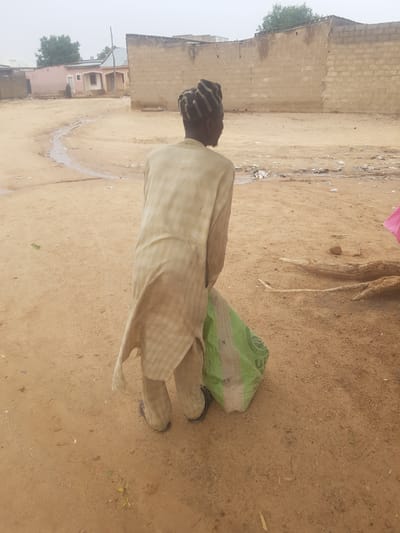 An elderly person is standing in a sandy area in Dutsinma B, Nigeria, near a dirt road and some buildings. The person is wearing a long, light-colored garment and a striped cap. A green bag is on the ground near the person's feet. The environment appears to be a residential area, with a block wall and other buildings visible in the background. There is also some debris scattered on the ground.